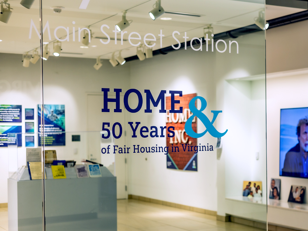 Photo of a portion of the 'HOME and 50 Years of Fair Housing' exhibit through the glass doors of a gallery.